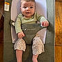 baby, infant, child, smiling, green_clothing, floral_pants, baby_seat, gray_fabric, mesh_backrest, wooden_floor, hands, feet, face, eyes, mouth, indoor, comfortable, cute, happy, sitting