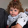 child, toddler, curly_hair, blue_eyes, white_sweatshirt, cartoon_print, indoor, couch, adult_in_background, person, portrait, reaching_hand, smiling, cute, home, family, relaxed, cozy, casual_clothing, living_room