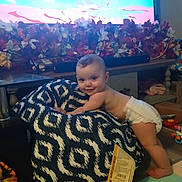 Mia is registered to the contest to win money with this photo: baby, diaper, bean_bag, smile, indoor, television, flower_arrangement, toy, floor_mat, child, pattern, cushion, furniture, play_area, home, cute, infant, person, happy, colorful