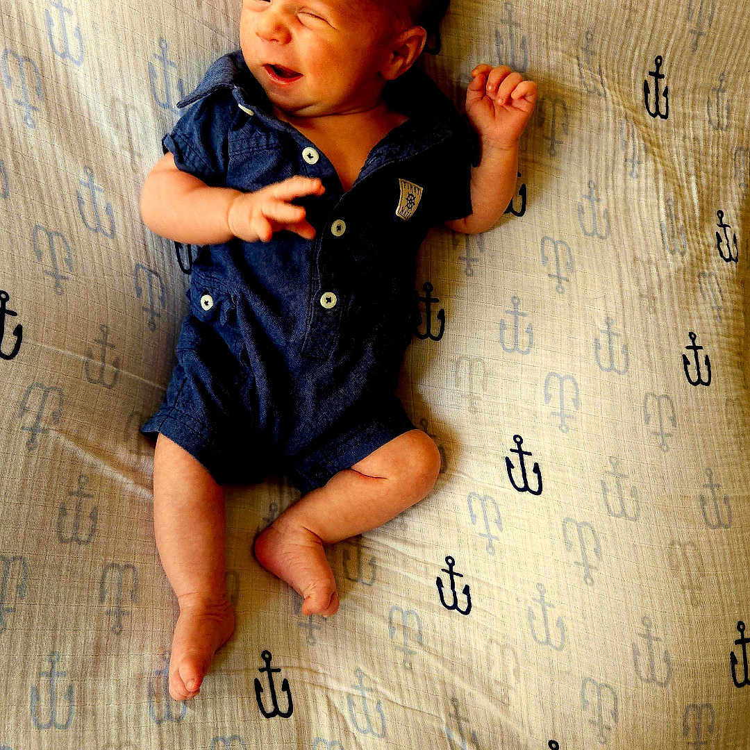 Silas is registered to the contest to win money with this photo: baby, infant, child, newborn, portrait, lying_down, winking, smiling, blue_outfit, onesie, mattress, bedding, indoor, closeup, face, hands, feet, brown_hair, patterned_background, cute
