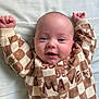 baby, infant, smiling, lying_down, checkered_clothing, onesie, bed_sheet, arms_up, close_up, portrait, cute, newborn, eyes_open, head, skin, cheeks, indoor, soft_lighting, blanket, patterned_fabric