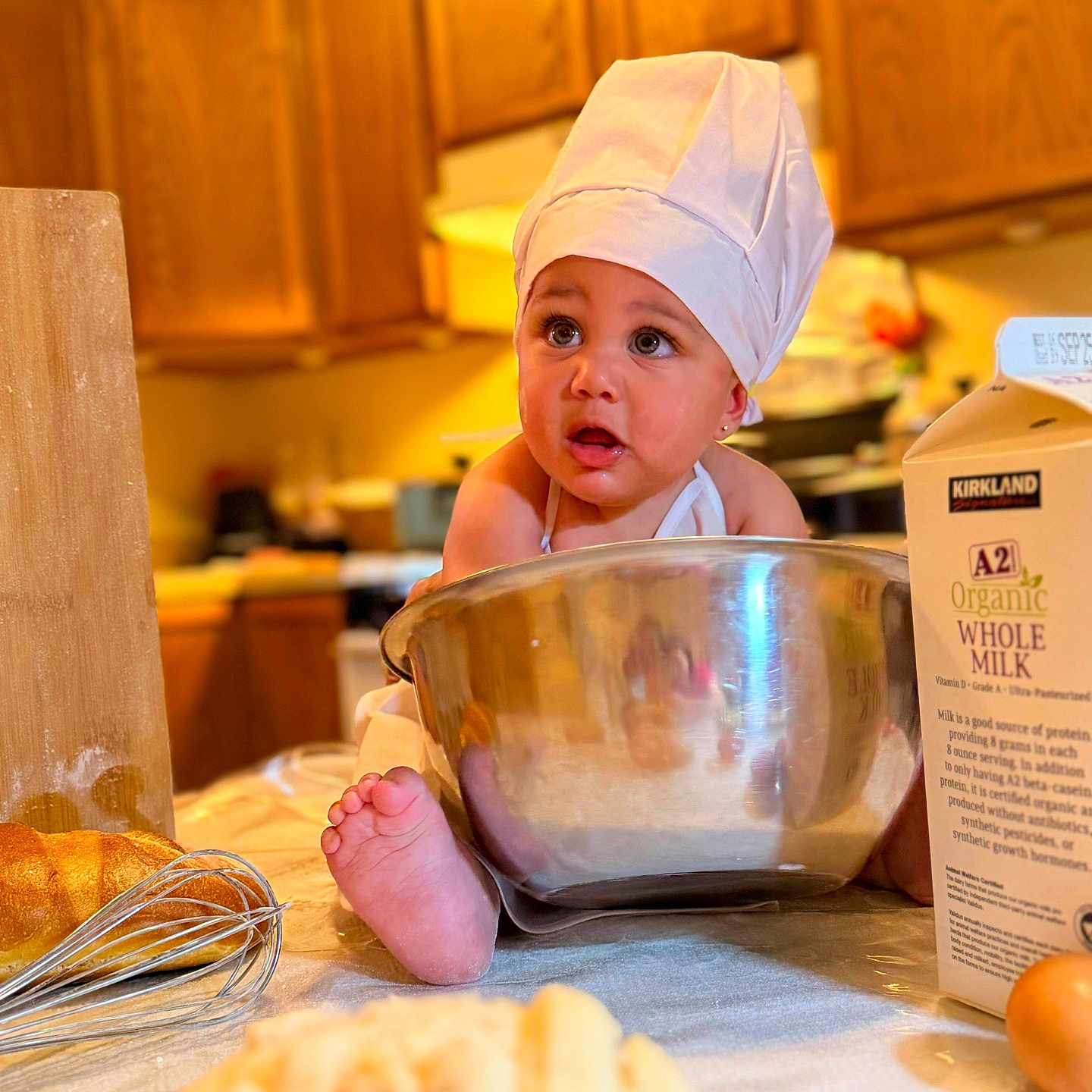 Indira joined the competition — help win amazing prizes! baby, child, chef_hat, mixing_bowl, kitchen, milk_carton, whisk, bread, countertop, wooden_cabinets, foot, table, cooking, baking, organic_milk, curious_expression, indoor, food_preparation, apron, ingredient