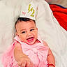 Indira is registered to the contest to win money with this photo: baby, child, smiling, pink_dress, crown, half_birthday, happy, cute, indoors, blanket, portrait, infant, closeup, celebration, accessory, bracelet, hand, foot, skin, person