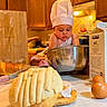 baby, chef_hat, baking, kitchen, mixing_bowl, bread, eggs, milk_carton, wooden_cabinets, countertop, apron, infant, food_preparation, cooking, child, indoor, candid, portrait, cute, domestic