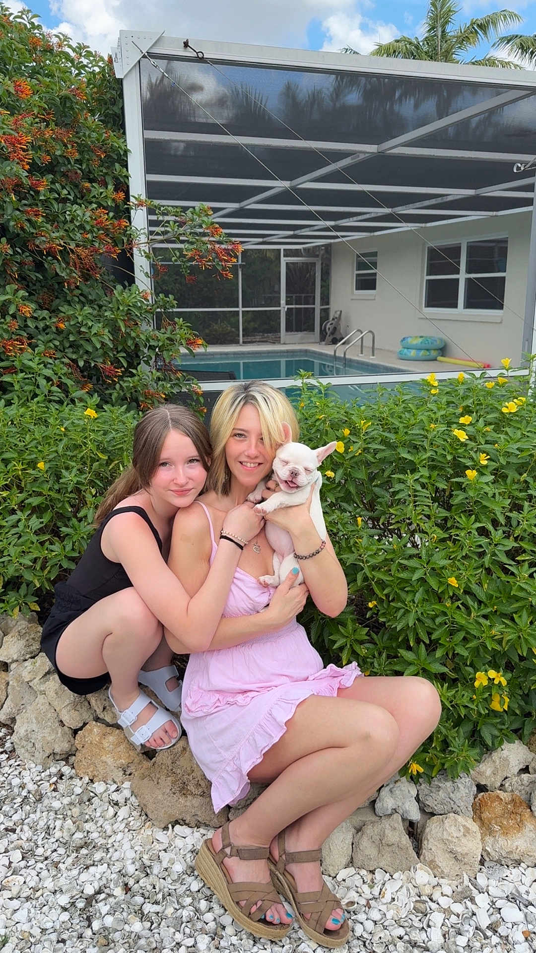Sophia Michelle Mae is registered to the contest to win money with this photo: two_women, dog, puppy, outdoor, garden, pool, smiling, friendship, plants, yellow_flowers, casual_clothing, summer, happy, crouching, sandals, rocks, leaves, sunny, nature, cute