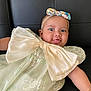baby, infant, child, bow, headband, dress, smiling, tongue_out, black_leather, couch, floral_pattern, cute, playful, happy, portrait, close_up, person, indoors, fashion, accessory