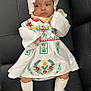 Yona is registered to the contest to win money with this photo: baby, infant, child, dress, embroidered, bonnet, white_clothing, socks, sitting, couch, black_cushion, cute, portrait, indoor, face, hand, leg, footwear, looking_away, curious