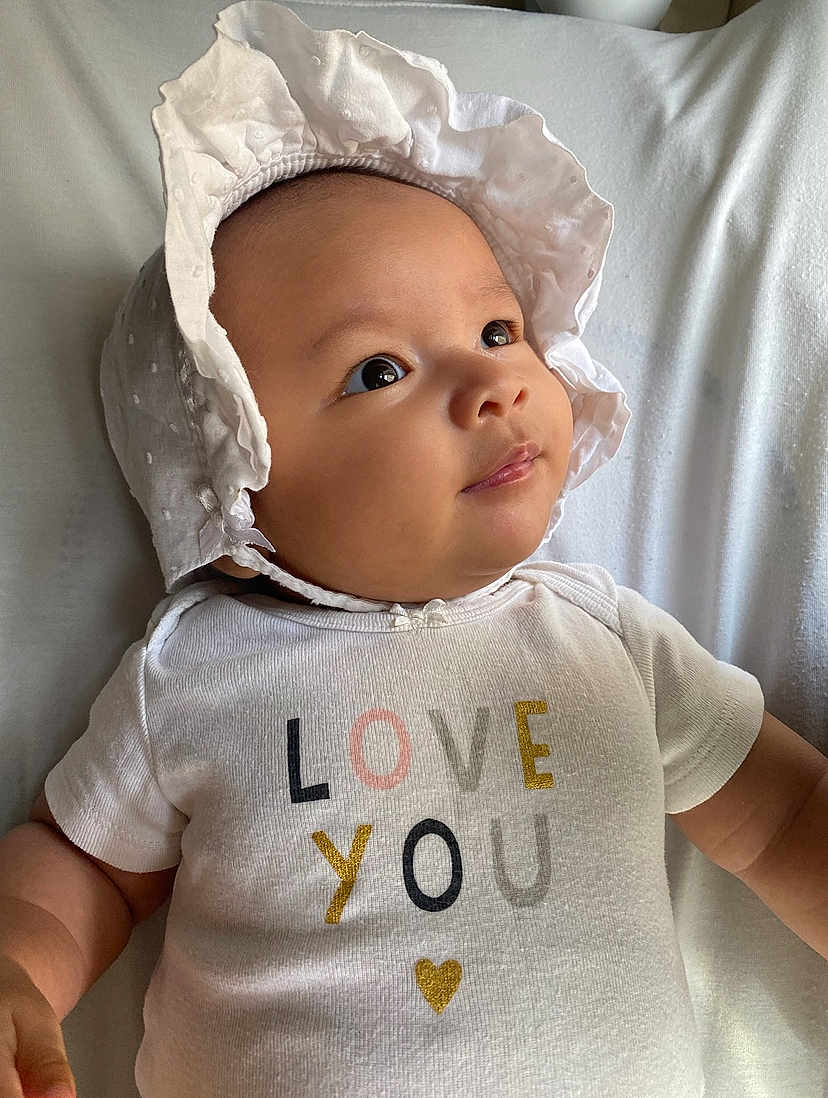 Yona is registered to the contest to win money with this photo: baby, infant, child, face, bonnet, white_clothing, text_on_clothing, love_you, lying_down, blanket, cute, portrait, soft_light, closeup, skin, expression, newborn, adorable, happy, indoor