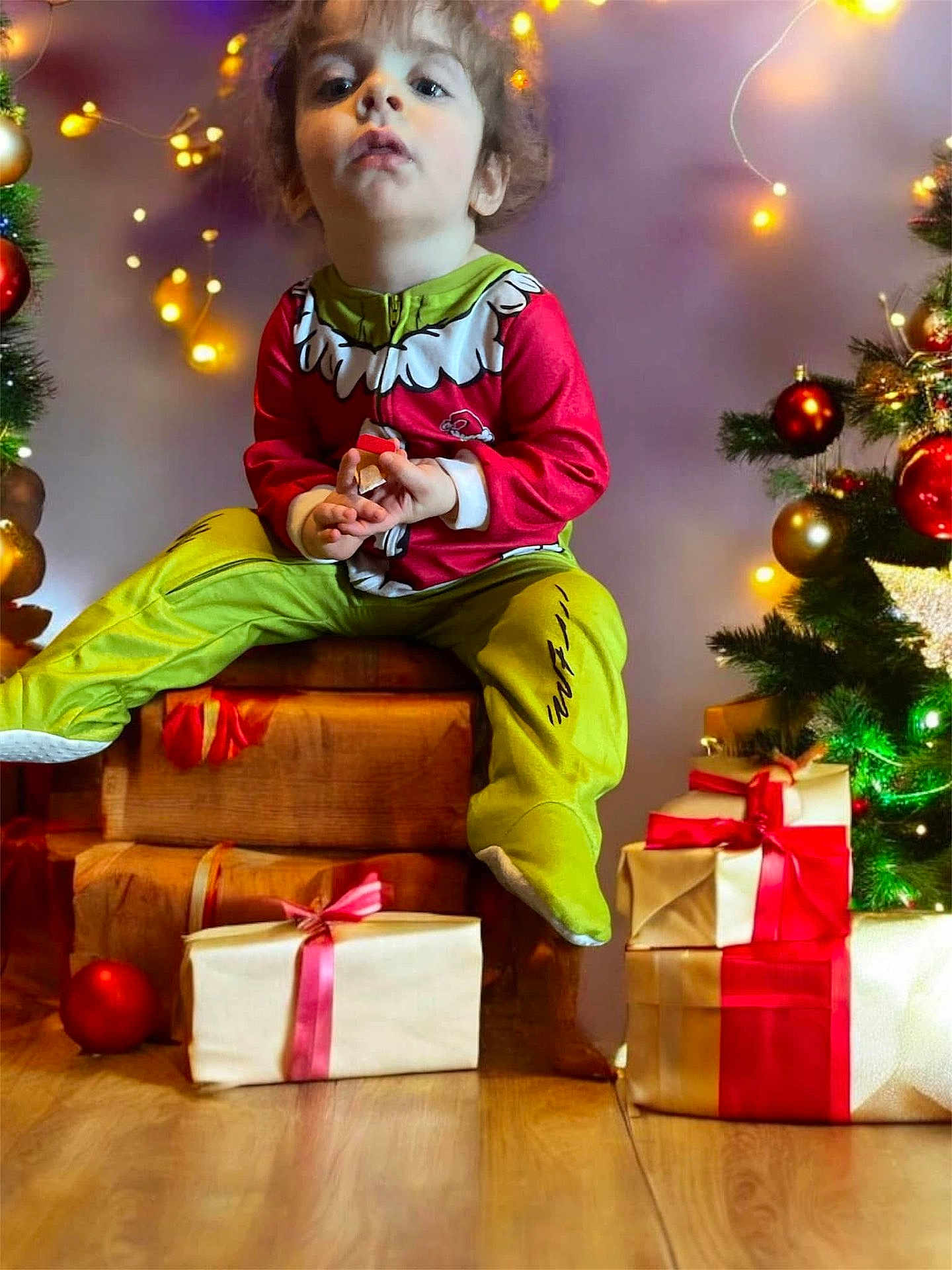 Kaden is registered to the contest to win money with this photo: child, toddler, holiday, christmas, pajamas, presents, gift, string_lights, decorations, tree, wooden_box, floor, indoors, festive, red, green, yellow, face, person, celebration