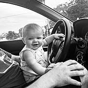 Briar is registered to the contest to win money with this photo: baby, child, car, steering_wheel, adult, hand, window, black_and_white, smile, shirt, seat, dashboard, keys, nature, trees, power_lines, interior, portrait, playful, happy