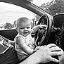 baby, child, car, steering_wheel, adult, hand, window, black_and_white, smile, shirt, seat, dashboard, keys, nature, trees, power_lines, interior, portrait, playful, happy