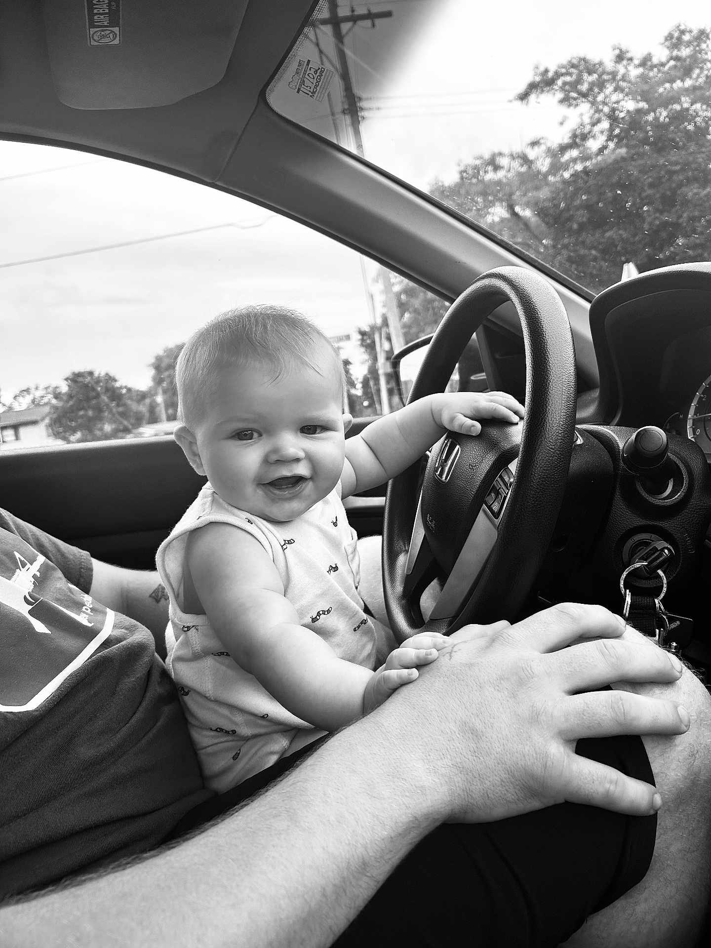 Briar is registered to the contest to win money with this photo: baby, child, car, steering_wheel, adult, hand, window, black_and_white, smile, shirt, seat, dashboard, keys, nature, trees, power_lines, interior, portrait, playful, happy