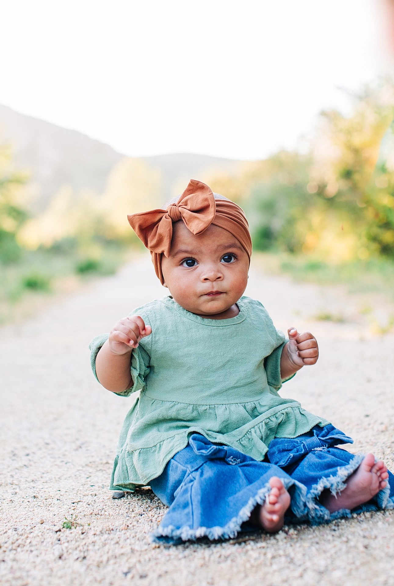 Daphne is registered to the contest to win money with this photo: baby, child, infant, headwrap, bow, green_dress, denim, jeans, barefoot, sitting, outdoor, gravel_path, portrait, bokeh, nature, trees, cute, big_eyes, expression, photography