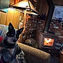 antler, blanket, bottle, brick_wall, couch, cozy, decor, dog, fan, fire, fireplace, furniture, home_interior, lamp, living_room, pet, sitting, warm_lighting, wood_paneling, wood_stove