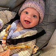 Aria-Maria is registered to the contest to win money with this photo: baby, smiling, knitted_hat, knitted_sweater, car_seat, pacifier, wooden_beads, happy, child, face, clothing, indoors, cute, person, seatbelt, infant, closeup, expression, accessory, warm_clothing