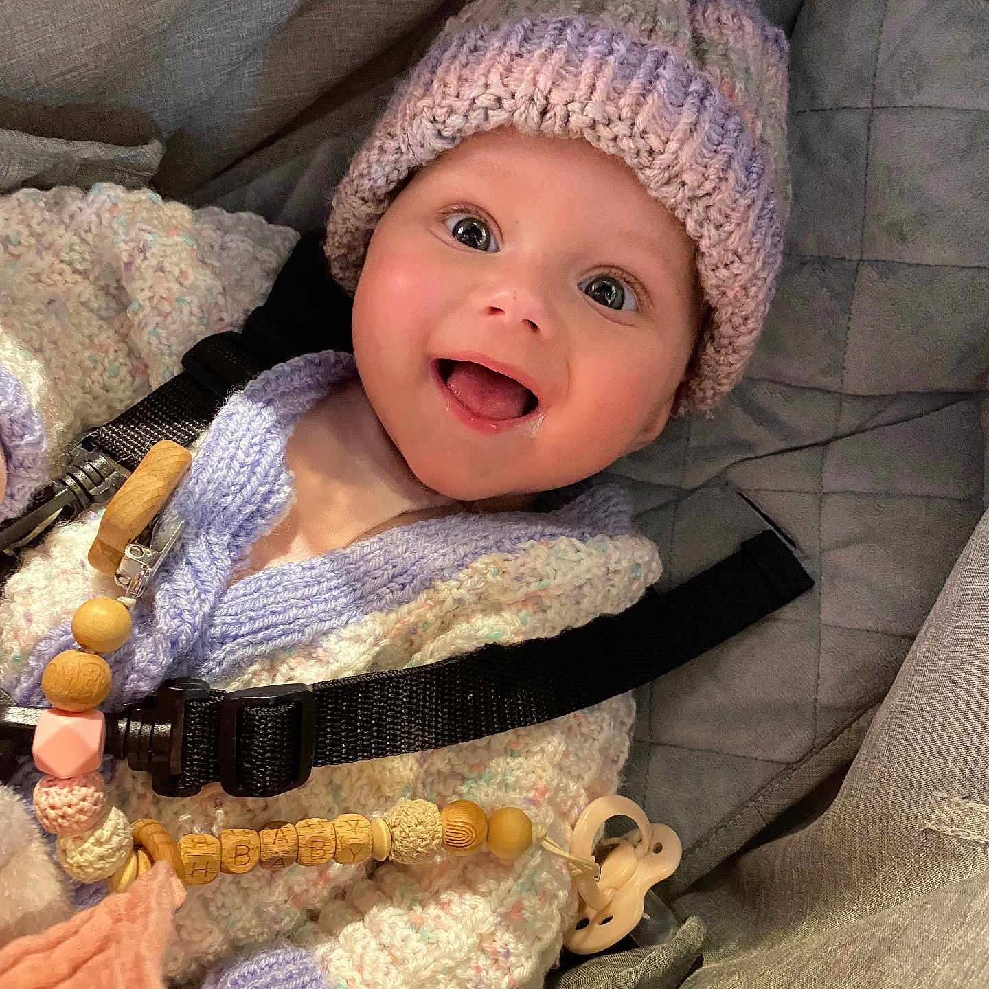 Aria-Maria is registered to the contest to win money with this photo: accessory, baby, car_seat, child, closeup, clothing, cute, expression, face, happy, indoors, infant, knitted_hat, knitted_sweater, pacifier, person, seatbelt, smiling, warm_clothing, wooden_beads