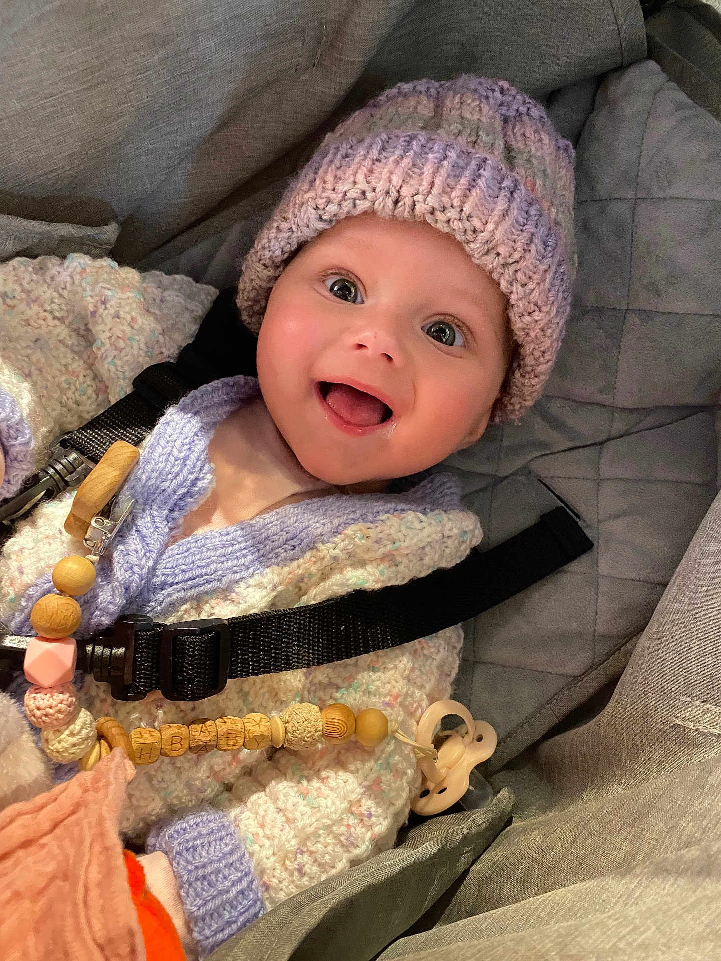 Aria-Maria is registered to the contest to win money with this photo: baby, smiling, knitted_hat, knitted_sweater, car_seat, pacifier, wooden_beads, happy, child, face, clothing, indoors, cute, person, seatbelt, infant, closeup, expression, accessory, warm_clothing