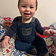 Akadian is registered to the contest to win money with this photo: baby, child, toddler, denim_overalls, smiling, happy, indoor, bed, bedspread, colorful, pattern, clothing, person, face, sitting, cute, young, infant, playful, joyful