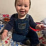 baby, child, toddler, denim_overalls, smiling, happy, indoor, bed, bedspread, colorful, pattern, clothing, person, face, sitting, cute, young, infant, playful, joyful