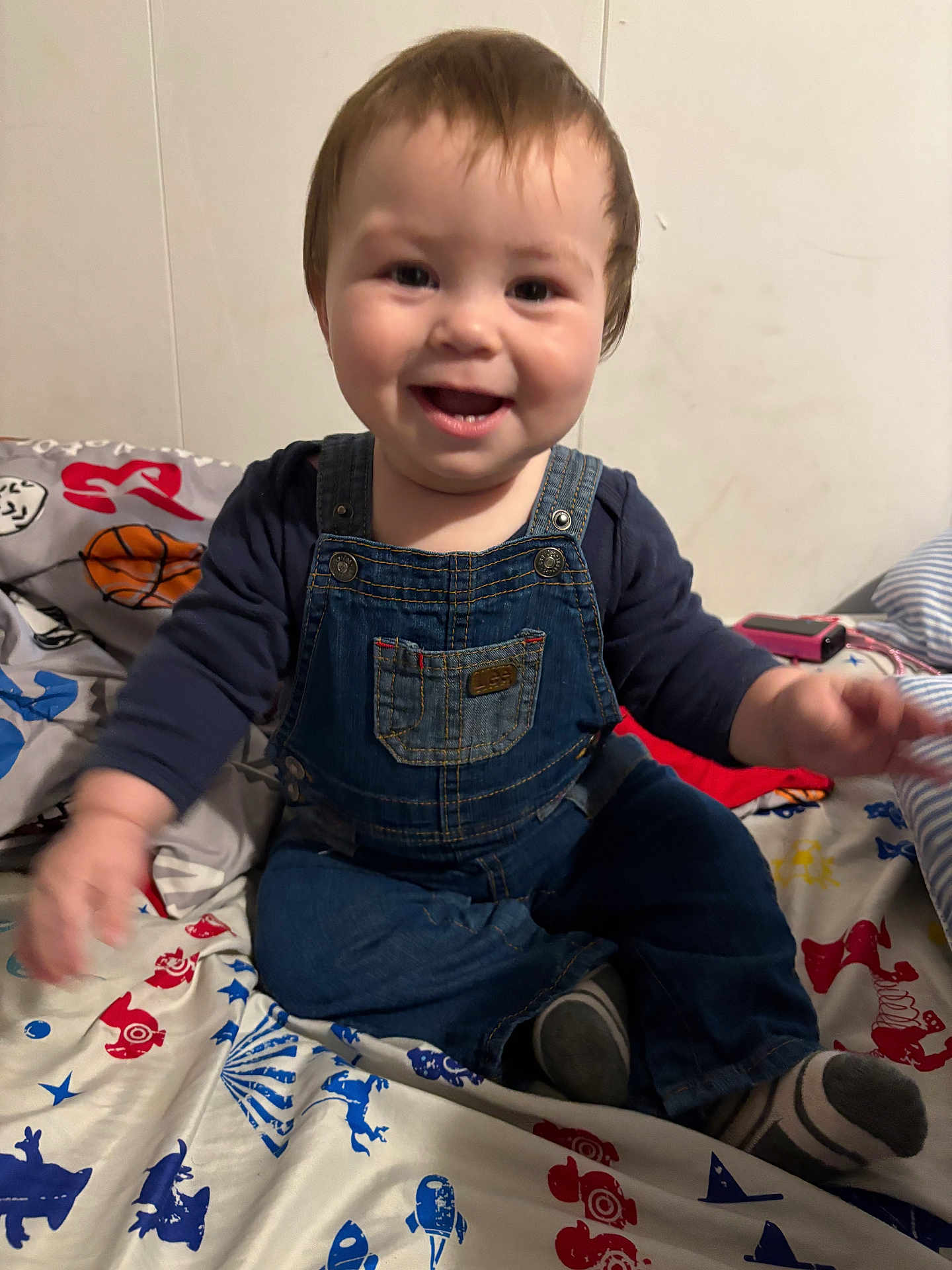 Akadian is registered to the contest to win money with this photo: baby, child, toddler, denim_overalls, smiling, happy, indoor, bed, bedspread, colorful, pattern, clothing, person, face, sitting, cute, young, infant, playful, joyful