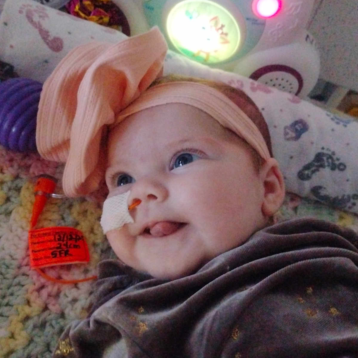 Azralina joined the competition — help win amazing prizes! baby, blanket, blue_eyes, bow, cheek, child, clothing, crochet, cute, face, feeding_tube, glowing, happy, headband, indoor, infant, lying_down, medical, smile, toy