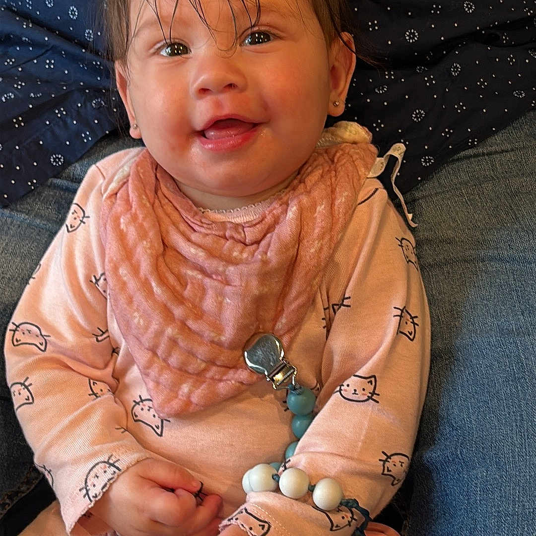 Evelyn joined the competition — help win amazing prizes! baby, child, infant, smiling, portrait, sitting, indoors, lap, adult, face, hair, eyes, pink_outfit, bib, pacifier, toy, clothing, couch, closeup, happy