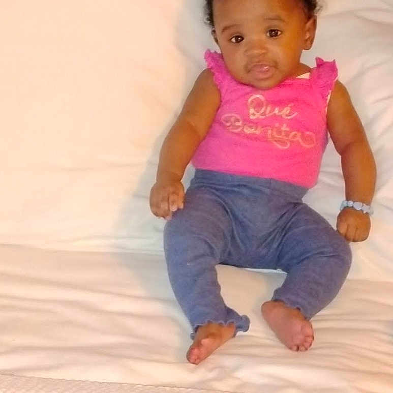 La'Daiyah joined the competition — help win amazing prizes! baby, child, girl, pink_bow, pink_top, blue_pants, bed, white_bedspread, pillow, wooden_headboard, indoor, portrait, sitting, cute, infant, person, clothing, footwear_free, bracelet, smiling
