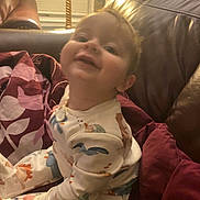 Miles is registered to the contest to win money with this photo: toddler, child, pajamas, smile, face, leather_couch, pillow, lamp, indoor, cozy, home, furniture, happy, portrait, baby, young_child, sitting, warm_lighting, blanket, casual