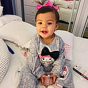 Adriana is registered to the contest to win money with this photo: toddler, child, bed, pillow, perfume_bottle, pink_bow, gray_outfit, cartoon_character, mirror, reflection, indoor, smiling, sitting, cute, hair_tied, footwear, soft_light, furniture, bedroom, person