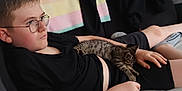 Orion a rejoint le concours — aidez-le/la à gagner de superbes lots ! black_shirt, blanket, boy, casual, cat, child, comfort, couch, glasses, home, indoor, kitten, pet, relaxed, resting, shorts, sleeping, soft_light, striped_cat, young