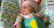 Addilyn is registered to the contest to win money with this photo: newborn, baby, infant, child, portrait, sitting, baby_seat, pink_hat, pajamas, floral_clothing, cushion, couch, indoor, home, colorful, patterned, awake, serious_expression, infant_toy, cozy