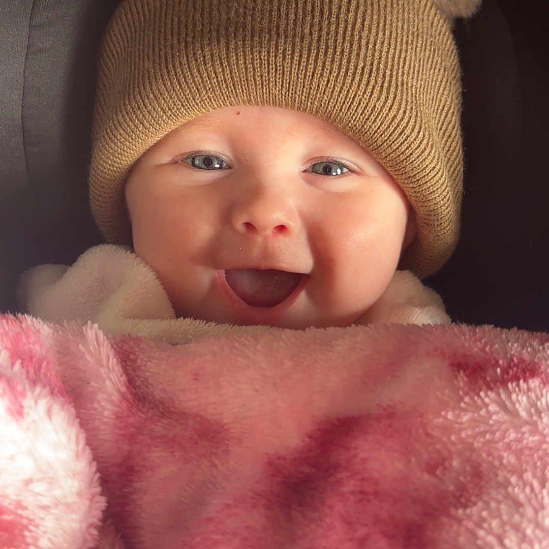 Mabel joined the competition — help win amazing prizes! baby, blanket, child, close_up, clothing, cozy, cute, face, happy, hat, headwear, indoor, infant, person, pink, pom_poms, portrait, smiling, soft, warm
