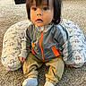 toddler, child, baby, sitting, carpet, pillow, dinosaur_pattern, jacket, pants, socks, indoor, floor, curious, face, person, young_child, clothing, hair, jewelry, home