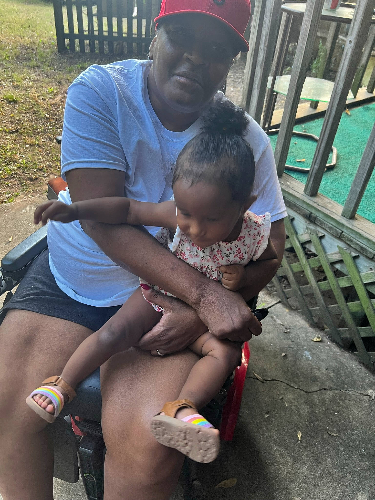 Keyanna is registered to the contest to win money with this photo: child, eyewear, fence, flip_flops, fun, hat, head, headwear, human_leg, interaction, knee, lap, leisure, person, recreation, sandal, shorts, sitting, t_shirt, thigh