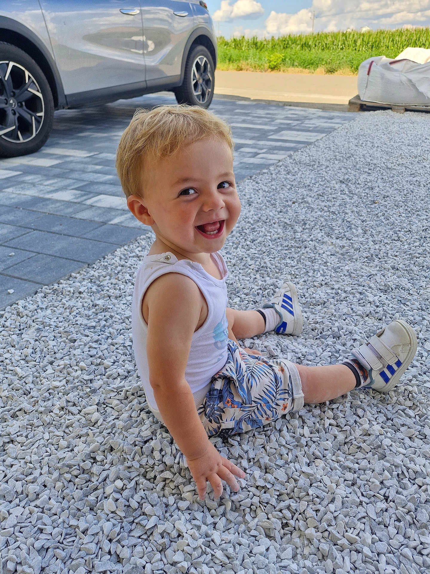 Elyam participe au concours pour gagner de l'argent avec cette photo : car, child, clouds, cute, daytime, grass, happy, nature, outdoor, pavement, person, shorts, sitting, sky, smiling, sneakers, stones, tank_top, toddler, young