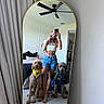 woman, mirror, selfie, kitten, dog, bandana, shirt, bedroom, bed, pillow, curtain, ceiling_fan, floor, tile_floor, shorts, crop_top, pet, indoor, reflection, phone