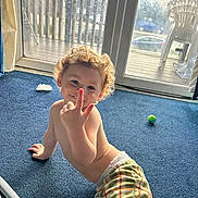 Albie is registered to the contest to win money with this photo: toddler, child, curly_hair, blue_carpet, plaid_pants, indoor, glass_door, smile, playful, bare_chest, toy_ball, sunlight, window, home, carpet_floor, cheeky, young_child, candid, curly_hair_child, happy