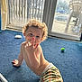 toddler, child, curly_hair, blue_carpet, plaid_pants, indoor, glass_door, smile, playful, bare_chest, toy_ball, sunlight, window, home, carpet_floor, cheeky, young_child, candid, curly_hair_child, happy