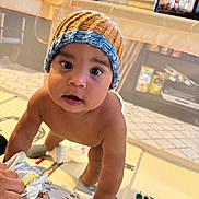 Ibrahim joined the competition — help win amazing prizes! baby, child, infant, portrait, knitted_hat, blue_hat, brown_hat, curious, wide_eyes, smiling, indoor, playpen, nursery, toys, blanket, adult_hand, sitting, leaning, family, home