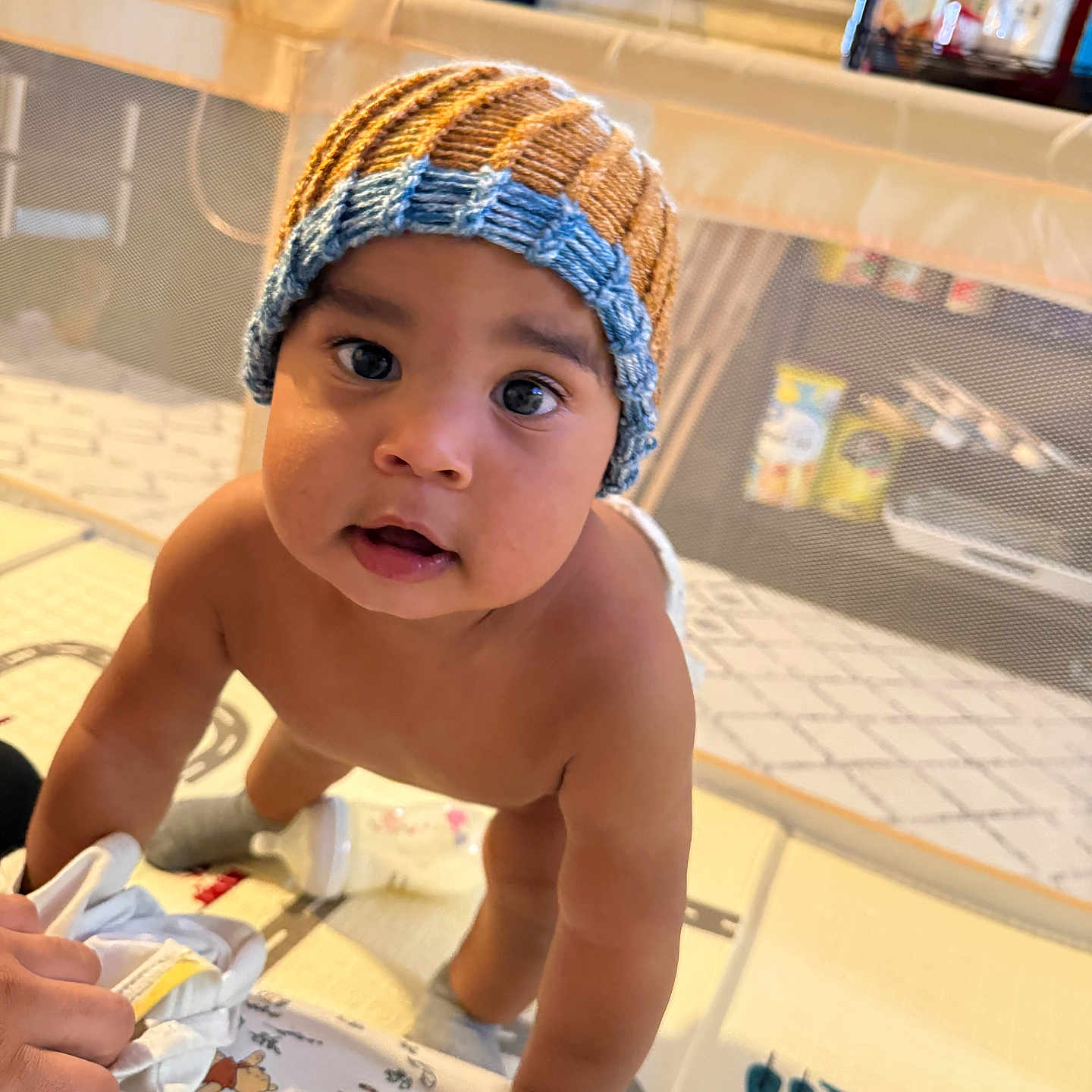 Ibrahim joined the competition — help win amazing prizes! baby, child, infant, portrait, knitted_hat, blue_hat, brown_hat, curious, wide_eyes, smiling, indoor, playpen, nursery, toys, blanket, adult_hand, sitting, leaning, family, home