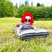 Ember is registered to the contest to win money with this photo: baby, child, sunglasses, red_hat, striped_pillow, grass, outdoor, field, summer, greenery, trees, nature, cute, sun_protection, infant, casual, daylight, fashion, portrait, relaxing