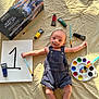 baby, infant, paint, acrylic_paint, paint_brush, palette, canvas, number_one, mustache, face_paint, denim_overalls, art_supplies, lying_down, indoors, wrinkled_sheet, colorful, creative, cute, small, expression
