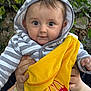 baby, child, hoodie, striped_clothing, yellow_fabric, hands, outdoor, greenery, tree_leaves, face, portrait, cute, person, smiling, holding, clothing, japanese_text, logo, closeup, infant