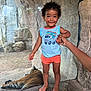 Tamãn is registered to the contest to win money with this photo: toddler, child, holding_hand, barefoot, tiger, animal, zoo, rock, enclosure, glass_barrier, curly_hair, smiling, casual_clothing, blue_shirt, orange_shorts, outdoor, nature, wildlife, person, hand