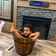 Tamãn is registered to the contest to win money with this photo: toddler, child, bucket, smiling, curly_hair, indoor, wooden_floor, fireplace, playful, happy, floor_vent, cabinet, wooden_bucket, black_clothing, home, flooring, face, feet, arms, person