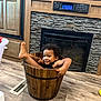 toddler, child, bucket, smiling, curly_hair, indoor, wooden_floor, fireplace, playful, happy, floor_vent, cabinet, wooden_bucket, black_clothing, home, flooring, face, feet, arms, person