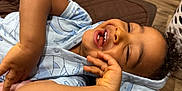 Tamãn is registered to the contest to win money with this photo: child, toddler, smiling, laughing, happy, curly_hair, blue_hoodie, whale_pattern, teeth, hand, face, lying_down, brown_quilt, wooden_floor, indoor, casual_clothing, cute, joyful, person, portrait