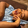child, toddler, smiling, laughing, happy, curly_hair, blue_hoodie, whale_pattern, teeth, hand, face, lying_down, brown_quilt, wooden_floor, indoor, casual_clothing, cute, joyful, person, portrait
