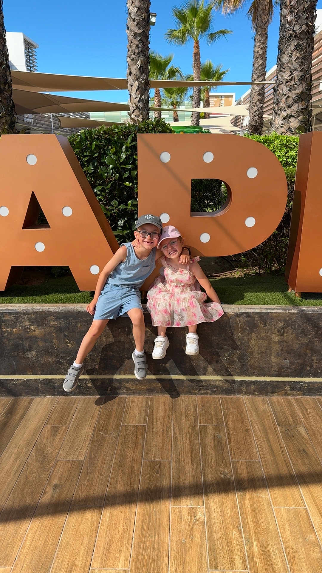 Tyler a rejoint le concours — aidez-le/la à gagner de superbes lots ! children, boy, girl, smiling, outdoor, daylight, palm_trees, large_letters, casual_clothing, glasses, hats, concrete_wall, wooden_floor, greenery, sunny, friendship, happy, posing, summer, playful