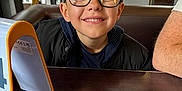 Tyler a rejoint le concours — aidez-le/la à gagner de superbes lots ! child, boy, glasses, smile, table, indoor, cafe, wooden_table, jacket, person, face, happy, reflection, casual, seated, short_hair, light, background, window, arm